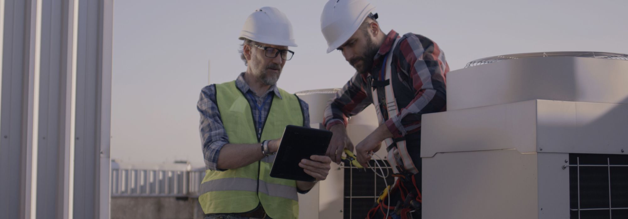 Two men standing on a rooftop near HVAC units while wearing safety hats and looking at an ipad.