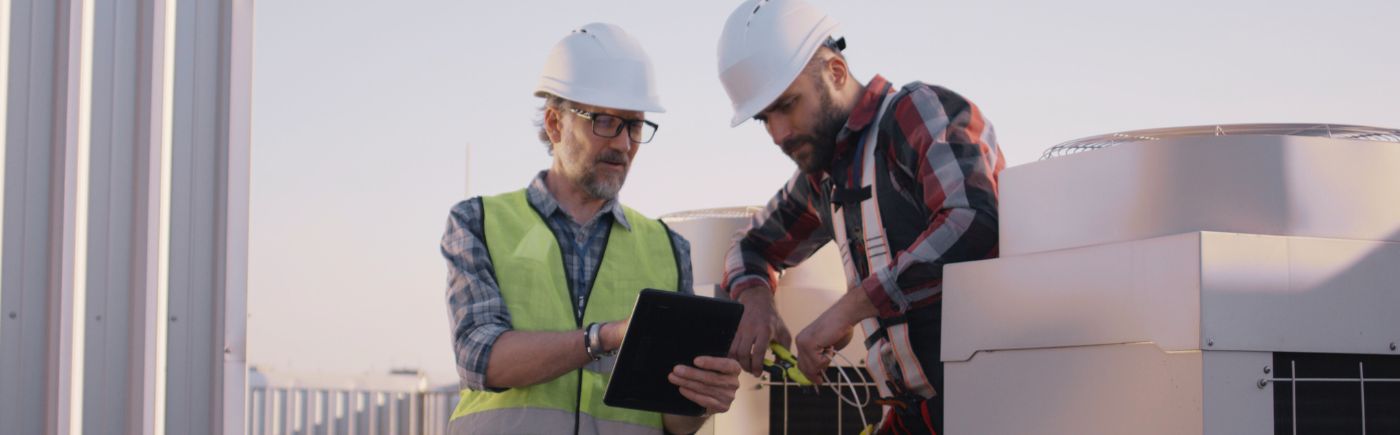 Two HVAC technicians wearing white hard hats look at an iPad screen. There is HVAC equipment in the photo.