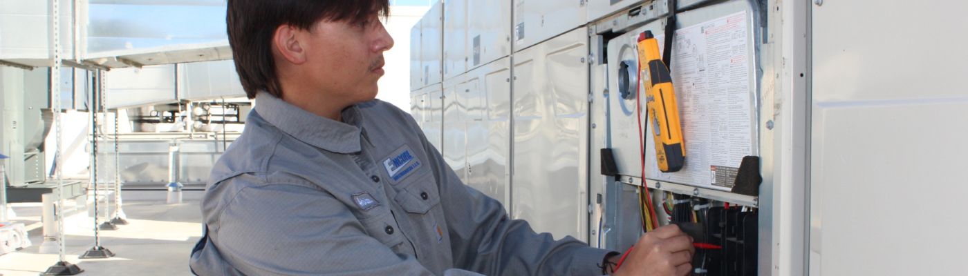 A close up of an HVAC technician wearing a gray shirt using tools on an rooftop HVAC unit.